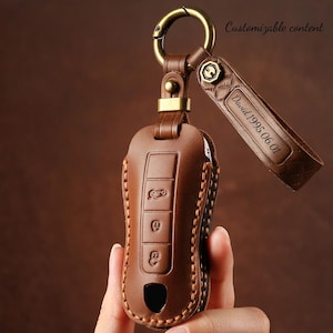 Puede incluir: Una funda de llavero de cuero marrón con un anillo y un cierre de metal dorado. La funda tiene tres botones y un logotipo de coche. Una correa de cuero está adjunta con el texto "David 1995.06.01". También se ve el texto "Customizable content".