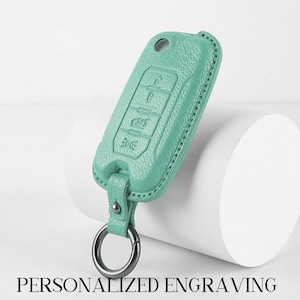 Puede incluir: Una funda de llavero de cuero verde azulado con un llavero plateado. La funda tiene símbolos en relieve para las funciones del coche. El texto "PERSONALIZED ENGRAVING" está en la parte inferior.