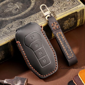 May include: Black leather car key fob cover with orange stitching and a matching keychain. The keychain has the text "Name 1995.06.01" and a metal clasp. The words "Customizable content" are also visible.
