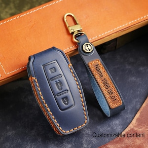 Puede incluir: Una funda para llave de coche de cuero azul marino con costuras naranjas y un llavero a juego. El llavero tiene una etiqueta de cuero con el texto "Name. 1995.06.01". Las palabras "Customizable content" están en la parte inferior.