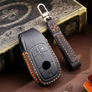 Puede incluir: Funda de llave de coche de cuero negro con costuras color canela y un llavero de cuero a juego. El llavero está grabado con el nombre "David" y la fecha "1995.06.01". La funda de la llave tiene un diseño de botones y un cierre metálico.