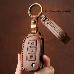 May include: A brown leather key fob case with a gold-colored metal clasp and a matching leather strap. The strap is engraved with the name "David" and the date "1995.06.01". The case has a stitched border and key buttons.