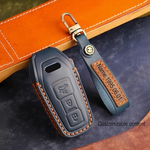 Puede incluir: Una funda para llave de coche de cuero azul marino y un llavero a juego con un cierre dorado. La funda de la llave tiene costuras naranjas y el llavero tiene el texto "Name.1995.06.01" en una etiqueta de cuero color canela. También es visible el texto "Customizable content".