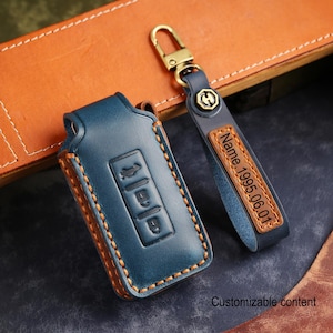 May include: A blue leather car key fob cover with visible brown stitching and a matching blue leather strap featuring a gold-toned metal clasp. The strap includes a rectangular patch with the engraved text Name, 1995.06.01 and Customizable content.