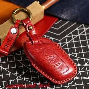 Puede incluir: Un llavero de cuero rojo con un llavero a juego. El llavero está grabado con símbolos de llaves de coche y cosido con hilo beige. Se adjunta una pequeña etiqueta roja con el nombre "GRACE". El llavero tiene un anillo color latón.