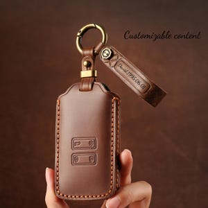 Op de afbeelding: Bruin lederen autosleutelhanger met een metalen sluiting en een klein leren label. Het label is gegraveerd met de tekst "David 1995 06 01". De sleutelhanger heeft een gestikte rand en reliëficonen. De tekst "Customizable content" is ook zichtbaar.