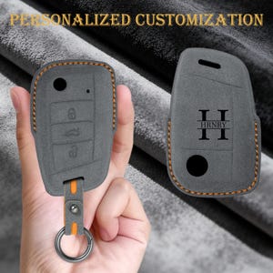 May include: Grey car key fob cover with orange stitching and a silver key ring. The cover has a personalised design with the initials "H R N R Y". The key fob cover has buttons for locking and unlocking the car.