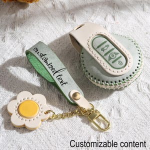 Puede incluir: Un juego de funda para llavero y llavero de cuero verde claro y crema. La funda del llavero tiene un diseño cosido y un cierre dorado. El llavero presenta un dije en forma de flor con un centro amarillo. El texto "Customized text" está en la correa.