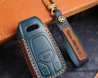 Könnte beinhalten: Eine blaue Autoschlüsselhülle aus Leder mit orangefarbenen Nähten und einem passenden blauen Schlüsselanhänger aus Leder. Das Band hat ein braunes Lederetikett mit der Prägung Name.1995.06.01 und einen messingfarbenen Metallverschluss.