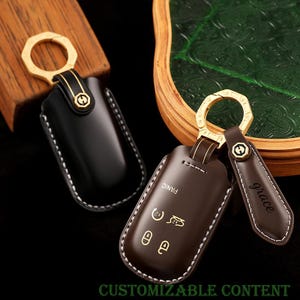 Könnte beinhalten: Zwei Leder-Schlüsselanhänger-Hüllen, eine schwarze und eine braune, mit goldfarbener Hardware. Die braune Hülle hat weiße Nähte und geprägte Symbole, mit dem Wort "panic" und dem Namen "grace" auf einem Anhänger. Der Text "CUSTOMIZABLE CONTENT" befindet sich unten.