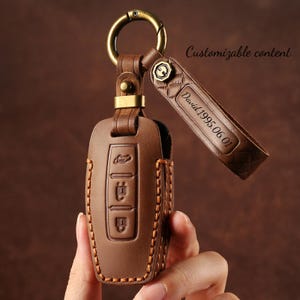 May include: Brown leather car key fob cover with a gold-colored keyring. The cover has three button icons and is stitched with orange thread. A leather tag is attached, engraved with "David 1995.06.01" and the words "Customizable content".