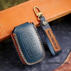 May include: A blue leather car key fob cover and matching keychain. The fob cover has embossed buttons and orange stitching. The keychain has a gold-colored clasp and a brown leather tag with the text "Name.1995.06.01".