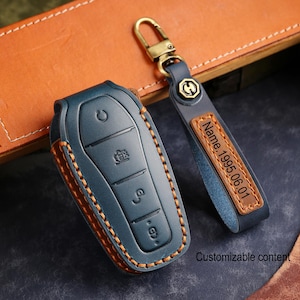 May include: A blue leather car key fob cover with orange stitching and a matching key chain. The key fob has three buttons and a key symbol. The key chain has a leather tag with the text "Name.1995.06.01".