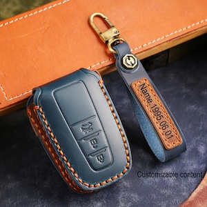 May include: A blue leather car key fob cover and matching keychain with a gold-colored clasp. The key fob cover has orange stitching and embossed buttons. The keychain has a leather tag with the text "Name.1995.06.01" and the words "Customizable content".