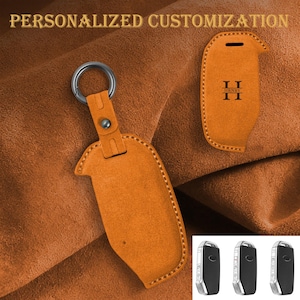May include: Orange leather key fob cover with a silver keyring. The cover has a stitched border and a snap closure. The text "PERSONALIZED CUSTOMIZATION" is at the top. A second cover is shown with the initials "H R N H Y".