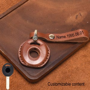 Può includere: Un portachiavi in pelle marrone con un design circolare e un cinturino. Il cinturino è stampato con "Name.1995.06.01". Il portachiavi ha una chiusura in metallo e un rivetto in metallo. Il testo "Customizable content" è in basso.
