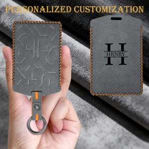 May include: A grey rectangular key fob holder with orange stitching and a metal ring. One side features a geometric pattern, while the other side displays the letter H and the text HRNRY. The holder includes a strap with a metal snap button.