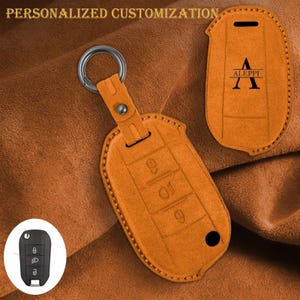 Pode incluir: Capa de chave de carro em couro laranja com um anel de metal e fecho de pressão. A capa tem símbolos em relevo para as funções da chave e o texto "ALEPPI" com a letra "A" em uma fonte estilizada. As palavras "PERSONALIZAÇÃO PERSONALIZADA" estão no topo.