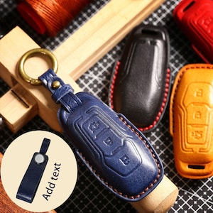 May include: A blue leather key fob cover with a gold-colored key ring. The cover has embossed buttons and stitching. Additional key fob covers in black, red, and orange are also visible.