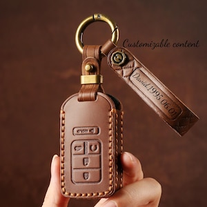 May include: A brown leather key fob case with a gold-colored metal ring and strap. The strap is engraved with "David 1995.06.01". The case has a stitched border and embossed key symbols.