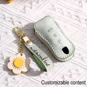Puede incluir: Un estuche para llave de coche de cuero verde claro y un juego de llavero a juego. La funda de la llave tiene costuras blancas y un ojo de cerradura negro. El llavero presenta un dije en forma de margarita con un centro amarillo y el texto "customized text".