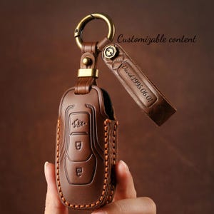 Peut inclure: Un étui de clé de voiture en cuir marron avec un porte-clés doré et une lanière. Le couvercle présente un motif cousu avec trois icônes de boutons. La lanière est gravée de "David 1995 06 01". Le texte "Customizable content" est également visible.