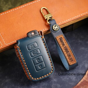 May include: A dark blue leather car key fob cover with visible orange stitching and a matching leather keychain strap. The strap features a rectangular brown leather patch embossed with the text Name. 1995.06.01 and a gold-toned metal clasp.