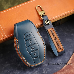 May include: A blue leather car key fob cover and matching key chain with gold-tone hardware. The key fob cover has embossed buttons and orange stitching. The key chain has the text "Name.1995.06.01" on a brown leather tag.