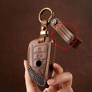 Puede incluir: Una funda de cuero marrón para la llave del coche con un llavero dorado. La funda tiene un diseño cosido con hilo negro y el texto "David 1995.06.01" es visible en la correa adjunta.