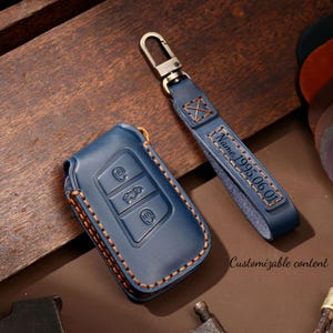 Puede incluir: Funda para llave de coche de cuero azul y llavero a juego. La funda de la llave tiene tres iconos de botones y está cosida con hilo marrón. El llavero tiene el texto "Name 1995.06.01" y un cierre metálico.