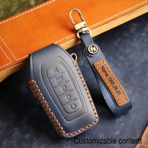May include: A navy blue leather car key fob cover with orange stitching and a matching key chain. The key fob cover has embossed buttons and the keychain has a leather tag with the text "Name.1995.06.01". The words "Customizable content" are at the bottom.