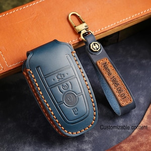 Puede incluir: Una funda para llave de coche de cuero azul y un llavero a juego con un cierre dorado. La funda de la llave tiene costuras naranjas y botones en relieve. El llavero tiene una etiqueta de cuero con el texto "Name.1995.06.01".