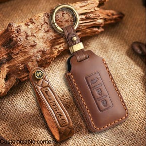 May include: Brown leather key fob cover with a gold-colored ring and a matching leather keychain. The key fob cover has embossed buttons and a stitched border. The keychain is engraved with the name "David 1995.06.01".