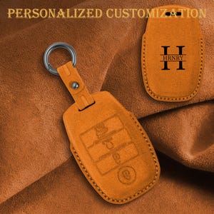 Könnte beinhalten: Eine hellbraune Autoschlüsselhülle aus Leder mit Metallring. Die Hülle zeigt geprägte Tasten-Symbole und ein separates Teil mit dem Buchstaben H, dem Namen HRNRY und dem Text PERSONALIZED CUSTOMIZATION in schwarzer Schrift.