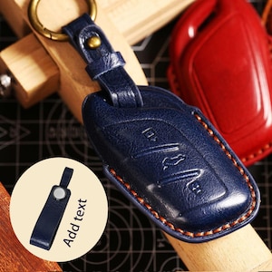 Puede incluir: Un llavero de cuero azul marino con un anillo dorado y una correa a juego. El llavero está cosido con hilo naranja y tiene detalles en relieve. Un llavero de cuero rojo está en el fondo. El texto "Add text" está en un círculo.