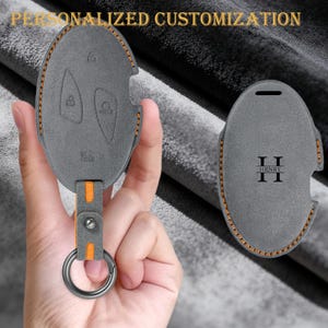 Puede incluir: Funda para llavero de cuero gris con costuras naranjas y una correa a juego. La funda presenta símbolos de llave en relieve y el texto "HOLD". La segunda funda tiene las iniciales "II" y el nombre "HRNRY" impresos. El texto "PERSONALIZACIÓN PERSONALIZADA" está en la parte superior.