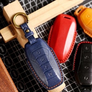 May include: A navy blue leather key fob cover with a brass-coloured ring and strap. The fob features embossed buttons and orange stitching. Other coloured key fob covers are also visible.