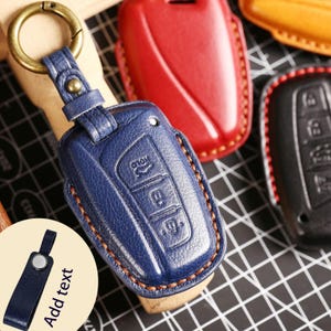 以下が含まれることがあります： 金色のキーリングが付いた、青い革製のキーフォブカバー。カバーにはエンボス加工されたボタンとステッチの縁取りがあります。赤、黒、黄色の他のキーフォブカバーが背景に見えます。画像には「Add text」というテキストが含まれています。
