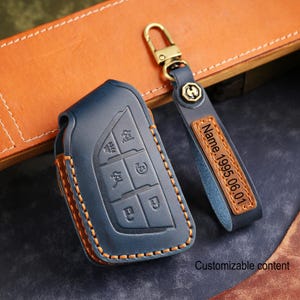 May include: A blue leather car key fob cover and matching keychain. The key fob cover has embossed buttons and orange stitching. The keychain has a leather tag with the text "Name.1995.06.01". The words "Customizable content" are also visible.