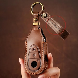 May include: A brown leather key fob with a gold-colored ring and accents. The fob has a stitched design and a rectangular tag with the engraved text "David 1995.06.01". The key fob is designed to hold a car key.