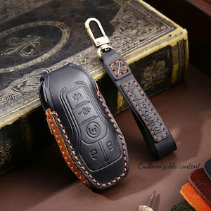 May include: Black leather key fob cover and matching keychain with gold-tone hardware. The key fob cover has a brown leather accent and visible buttons. The keychain is engraved with "David 1995.06.01". The words "Customizable content" are also visible.