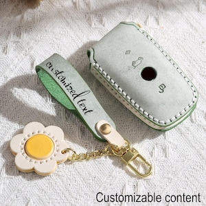 May include: A light green leather key fob case and matching key chain with a daisy charm. The key fob case has a stitched border and a car symbol. The key chain has a strap with the words "customized text".