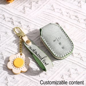 May include: A light green leather key fob cover and a matching keychain with a flower-shaped charm. The keychain has a gold-coloured clasp and chain. The key fob cover has white stitching and the words "customized text" on the strap.