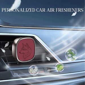 Puede incluir: Un ambientador de coche plateado sujeto a una rejilla de ventilación, con una cara cuadrada de color rojo oscuro y una ilustración de línea negra de una cabeza de lobo. El texto PERSONALIZED CAR AIR FRESHENERS aparece arriba, junto a burbujas con flores y limas.