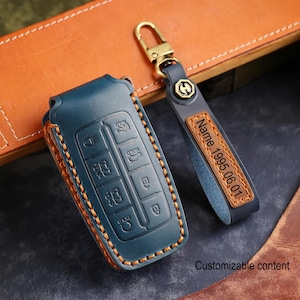 May include: A blue leather car key fob cover with orange stitching and a matching keychain. The keychain has a gold-coloured clasp and a leather tag with the text "Name 1995.06.01". The words "Customizable content" are also visible.
