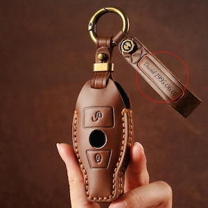Pode incluir: Um porta-chaves de couro castanho com um anel e ferragens douradas. O porta-chaves tem um design costurado e apresenta três botões. Uma etiqueta de couro está anexada, com o texto "David 1995.06.01" gravado.