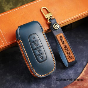 May include: A navy blue leather car key fob cover and matching keychain. The key fob cover has orange stitching and embossed buttons. The keychain has a gold-colored clasp and a leather tag with the text "Name.1995.06.01".
