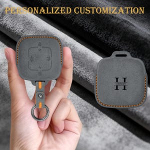 Peut inclure: Housse de clé de voiture grise avec des accents orange et un porte-clés en métal. La housse présente des symboles en relief et le texte "HRNRY". L'image comprend également le texte "PERSONNALISATION PERSONNALISÉE".