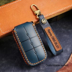 Puede incluir: Un estuche para llavero de cuero color turquesa y un llavero a juego con herrajes dorados. El estuche para llavero tiene costuras naranjas y botones en relieve. El llavero tiene una etiqueta de cuero con el texto "Name.1995.06.01". También es visible el texto "Customizable content".