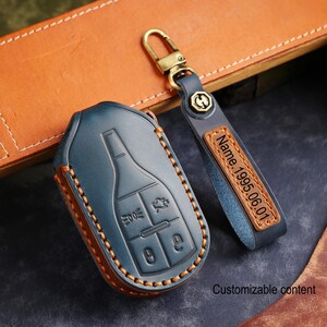 May include: A blue leather car key fob cover with orange stitching and a matching keychain. The keychain has a leather tag with the engraved text "Name.1995.06.01". The key fob cover has a car key design.